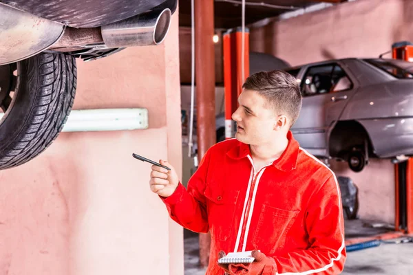 Male motor mechanic standing making notes underneath a lifted ca — Stock Photo, Image