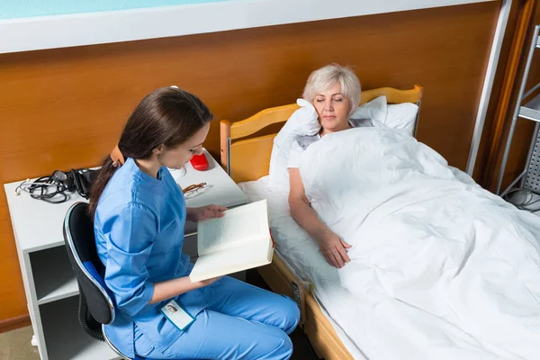 Young nurse in uniform is reading a book for her patient — Stock Photo, Image