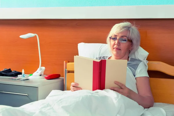 Patientin mit Brille liest ein Buch, während sie im Bett liegt — Stockfoto