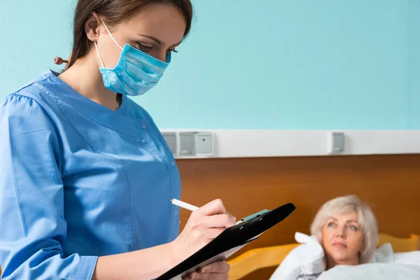 Attraente infermiera in uniforme indossando una maschera facendo alcune note in — Foto Stock