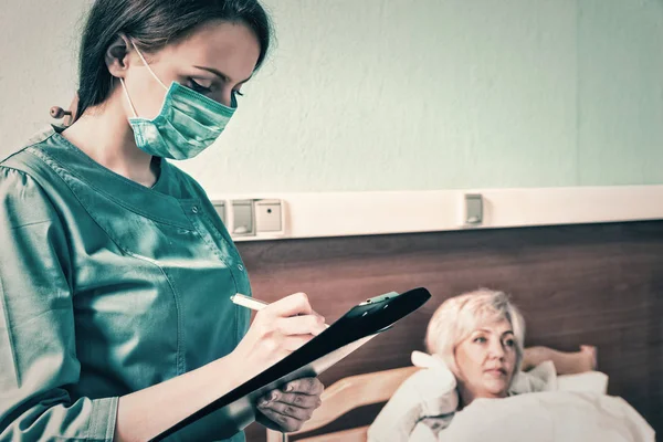 Giovane infermiera in uniforme indossando una maschera facendo alcune note in repor — Foto Stock