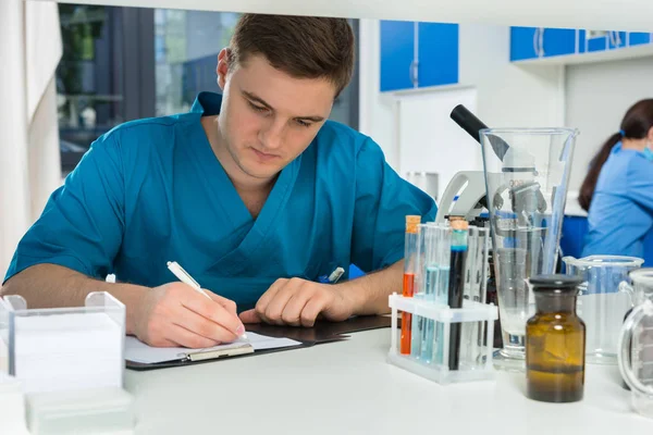 Young male scientist in uniform writing down notes of his resear — Stock Photo, Image
