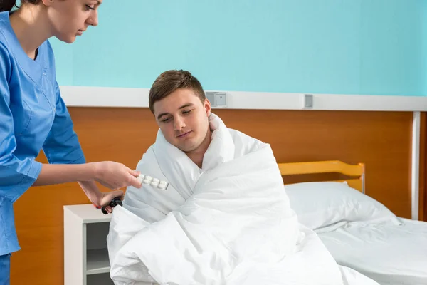 Enfermera joven dando pastillas al paciente enfermo sentado en silla de ruedas co —  Fotos de Stock