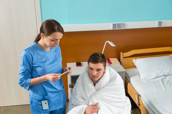 Молодая медсестра в форме смотрит на термометр пациента, который — стоковое фото