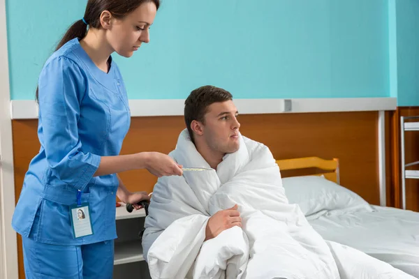 Sygeplejerske i uniform kigger på et termometer af patient, der sidder - Stock-foto
