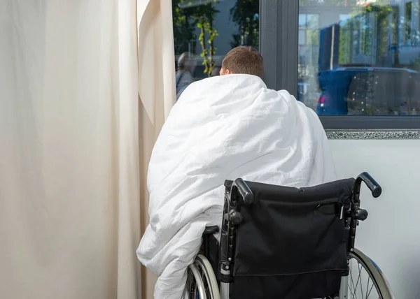 Нездоровый пациент, сидящий у окна на инвалидной коляске, покрытый qui — стоковое фото