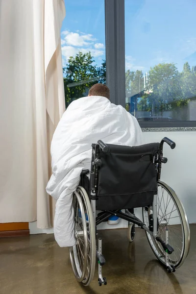 Männliche junge Patientin sitzt am Fenster auf Rollstuhl bedeckt w — Stockfoto