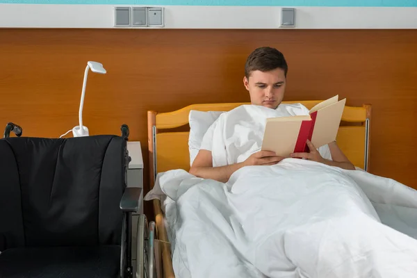 Paciente varón joven enfermo acostado en la cama del hospital y leyendo un b — Foto de Stock