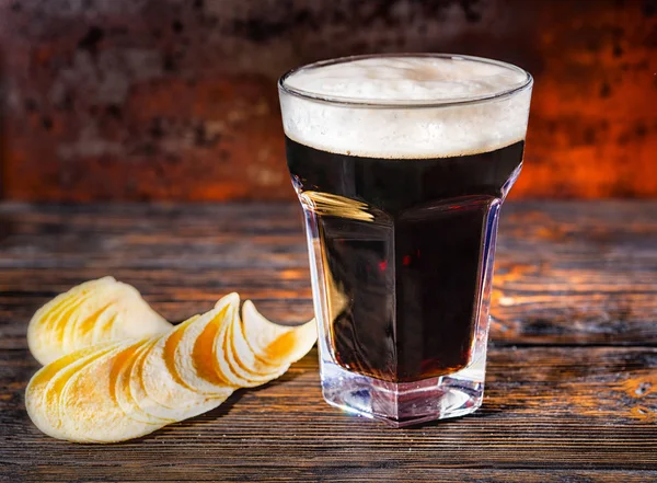 Стакан свежего тёмного пива рядом с чипсами на тёмном деревянном столе — стоковое фото