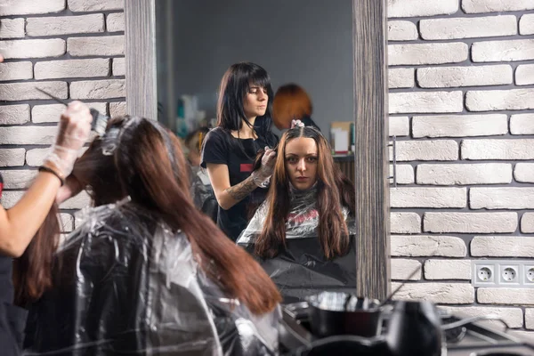 Reflection in large mirror of female stylist stands and dyeing h