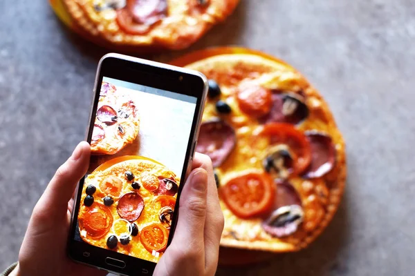 Teenage girl`s hands with smartphone takes picture of fresh just baked hot self-made pizza with salami, tomato, cheese, olives and mushrooms on grey grunge table background.