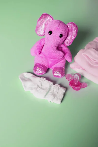 Cute pink toy elephant, infant clothes and pacifier on light green background with copy space for text or message  as template of greeting card for new parents. Concept of waiting for baby.
