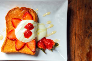 Yoğurtlu tost, beyaz tabakta çilek ve bal, ahşap masada kahvaltı.