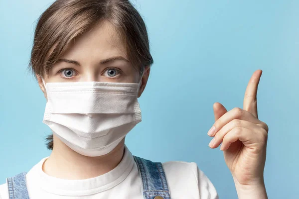 Beautiful girl in a white medical mask shows thumb up, blue background
