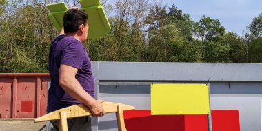 Şişman, koyu saçlı, orta yaşlı bir adam, güneşli bir günde Almanya, Avrupa 'da büyük boy çöp toplama istasyonundaki büyük konteynırlara ahşap tahtalar ve çöp atmak için çöp taşır..