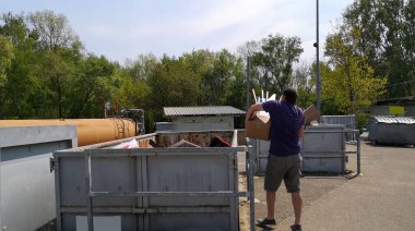 Şişman, koyu saçlı, orta yaşlı bir adam, güneşli bir günde Almanya, Avrupa 'daki büyük boy çöp toplama istasyonunda, içi ev atığı dolu karton kutuları büyük bir konteynıra atıyor..