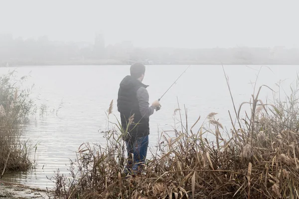 Pêcheur homme pêche avec canne à filer sur une rive de la rivière au lever du soleil brumeux brouillard — Photo