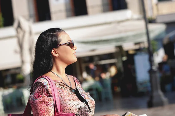 Женщина в солнечных очках, держит карту, идет по улице со старыми зданиями и смотрит в сторону — стоковое фото