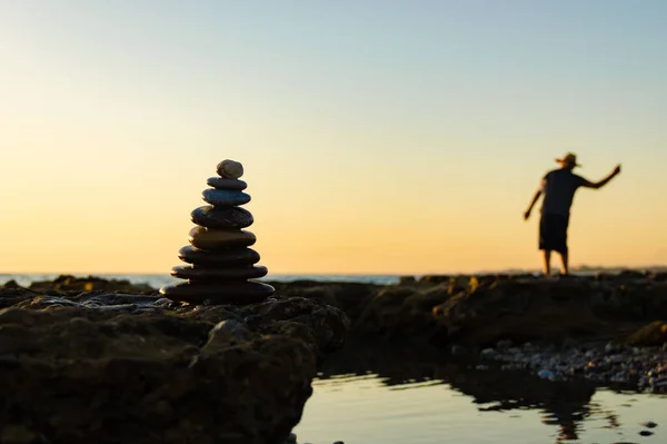 Silhueta de uma pirâmide de seixos e um homem desfocado jogando pedras no mar . — Fotografia de Stock