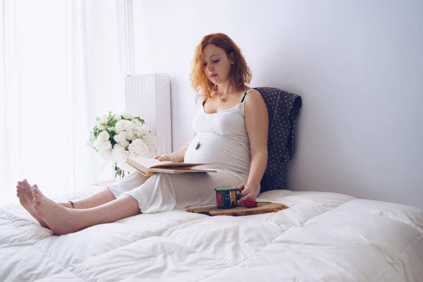 Giovane donna incinta rilassante sul letto con un libro — Foto Stock
