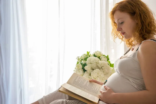 Schwangere berührt ihren Bauch, während sie ein Buch liest — Stockfoto