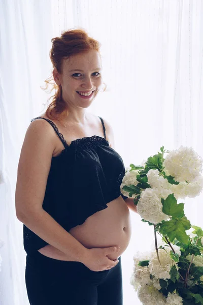 Jonge mooie zwangere vrouw staat in de buurt van raam thuis — Stockfoto