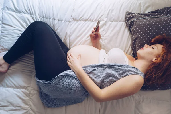 Zwangere vrouw liegt in bed met smartphone, bovenaanzicht — Stockfoto