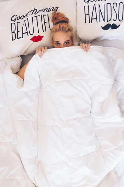 Ung kvinna ligger i sängen medan döljer hennes ansikte under täcket — Stockfoto