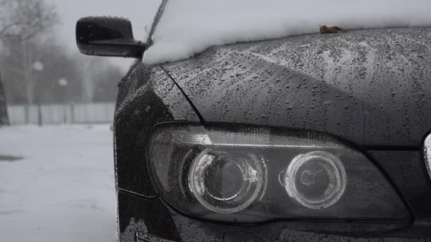 A headlamp closeup during snowfall in winter daytime. Bad weather conditions for traffic, blizzard. Danger for trips. Glassy black ice on the ground road. Snow covered automobile is in yard near house — Stock Video