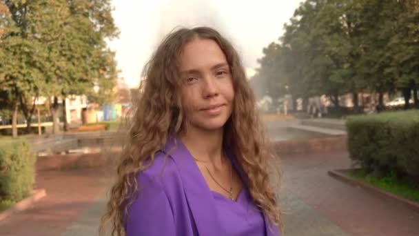 Video Retrato de una hermosa chica con el pelo rizado sobre un fondo de la ciudad, de cerca. Linda chica con una chaqueta púrpura sonríe a la cámara, cámara lenta — Vídeos de Stock