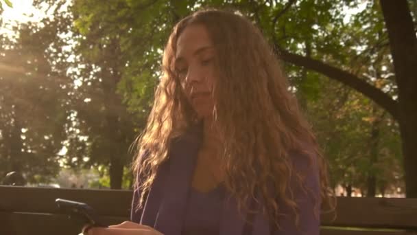 Caucásico chica con el pelo largo se sienta en un banco en el parque y navega en el teléfono Internet, fondo, lento mo — Vídeos de Stock