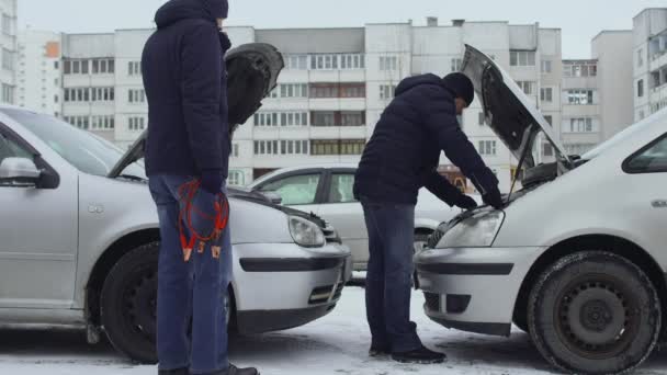 Men are going to charge discharged automotive battery with jumper clamp cables connecting them to post terminal. Hoods raised up in winter daytime in parking. Malfunction or problem with auto. Part 3 — Stock Video