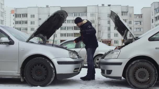 O homem conecta cabos de braçadeira de jumper da bateria automotiva descarregada ao terminal de postagem da bateria. Capuzes levantadas durante o dia de inverno no estacionamento do quintal. Mau funcionamento ou problema com o automóvel. Parte 3 — Vídeo de Stock