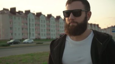 Siyah gözlüklü, gür sakallı, yakışıklı, beyaz bir adam havalarda ve arka planda yürüyor. Güneşli gün batımı, yavaş çekim, model