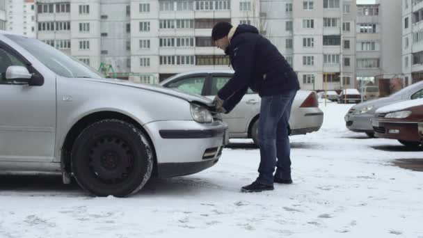 Мужчина пытается и не может открыть капот своей машины зимой на парковке двора. Капот замерз из-за холодной погоды. Неисправность или проблема с автоматикой. Срыв. Хочется зарядить батарею — стоковое видео