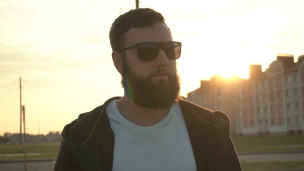 Un hombre caucásico joven guapo con una barba tupida en gafas negras camina en el clima, fondo. Puesta de sol soleada, cámara lenta, modelo — Vídeo de stock
