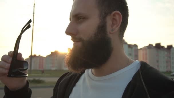 Knappe jonge blanke man model zet op zwarte bril op zonsondergang achtergrond. Mooie bossige baard op een achtergrond van de zon, portret, slow motion — Stockvideo