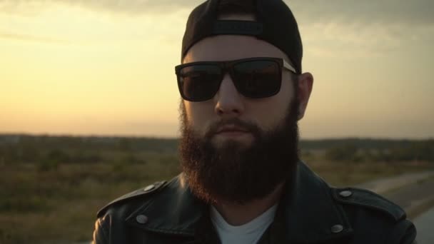 Un hombre guapo y brutal con barba gruesa y gafas negras se levanta sobre el fondo de una puesta de sol con los brazos cruzados. Mirando a la cámara, cámara lenta — Vídeo de stock