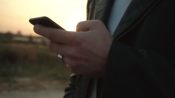 Um homem corre o dedo na tela de um smartphone contra o pano de fundo de um pôr do sol ensolarado, close-up. Fundo, câmera lenta — Vídeo de Stock