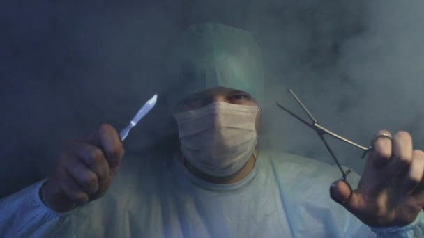 Хирург со скальпелем, кузнечиком или щипцом смотрит на камеру в дыму в замедленной съемке. Врач готов начать резать ткани пациентов. Спасти жизнь или убить концепцию. Операционный стол и операционный стол — стоковое видео