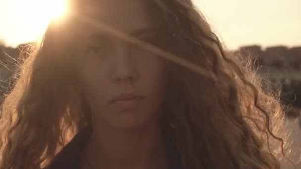 Young beautiful girl with long curly hair raises her eyes and looks at the camera. Against the background of a sunny sunset, close-up, video portrait — Stock Video