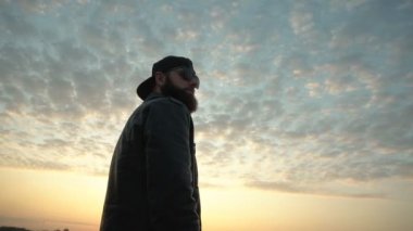 Siyah gözlüklü, yakışıklı beyaz adam şehirde güneşli bir günbatımının arka planına bakıyor. Arka planda. Yavaş çekim