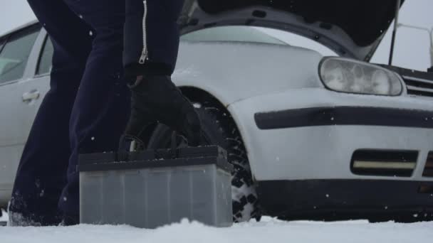 O homem levanta a bateria automotiva do chão coberto de neve e carrega seu carro no inverno. Um repara ou corrige o carro quebrado na queda de neve. Avaria ou problema com o veículo. Capuz ou capota levantada — Vídeo de Stock