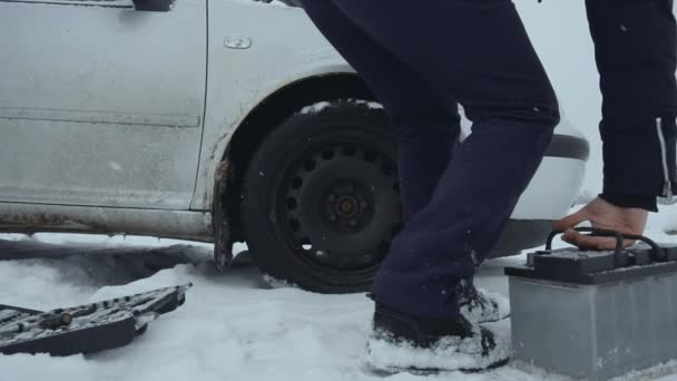 O homem levanta a bateria automotiva do chão coberto de neve e carrega seu carro no inverno. Um repara ou corrige o carro quebrado na queda de neve. Avaria ou problema com o veículo. Capuz ou capota levantada — Vídeo de Stock