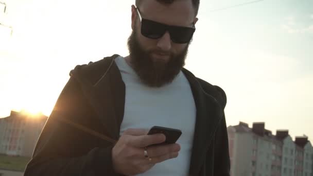 O tipo caucasiano jovem na moda com uma barba e em copos pretos comunica-se na Internet no telefone contra o pôr-do-sol, câmera lenta — Vídeo de Stock