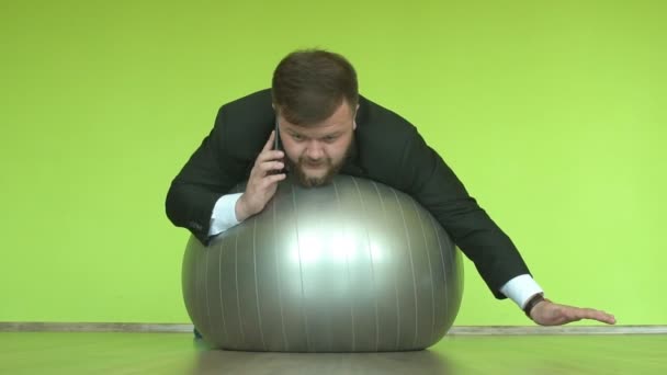 Красивый белый мужчина с бородой в офисном костюме разговаривает по телефону и качается на фитнес-мяче, медленно — стоковое видео