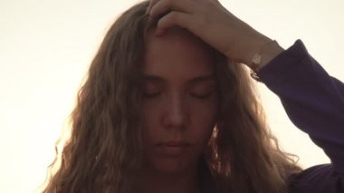 Uzun kıvırcık saçlı genç güzel kız gözlerini kaldırır ve kameraya bakar. Güneşli bir günbatımının arka planında, yakın plan, video portresi