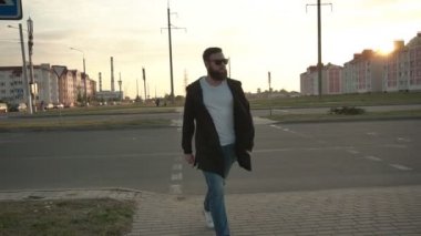 Siyah gözlüklü yakışıklı beyaz adam, sakallı, gün batımına karşı şehirde yürüyor, ağır çekimde. Şık ve şık bir adam..