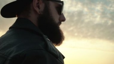 Çalı sakallı yakışıklı vahşi bir adam güneşli turuncu bir gün batımının arka planında yürüyor. Siyah gözlüklü genç adam ve düz vizörlü bir şapka, kopyalama alanı, yavaş çekim, Kafkas