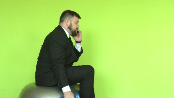 一个身穿黑色西服、留着胡子的高加索男性商人坐在一个健身球上，带着哑铃走进去。 讲电话和运动，复制空间 — 图库视频影像